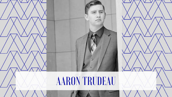 A professional-looking Black-and-white portrait of a young man in a suit and tie, facing slightly left with a confident and composed expression. The geometric patterned background is overlaid with a semi-transparent white band at the bottom.